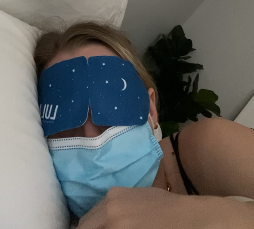 Selfie image of Annie in bed asleep. She is wearing a paper eye mask over her eyes, and a surgical mask over her mouth and nose.