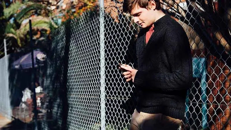 Young male leaning on fence using phone