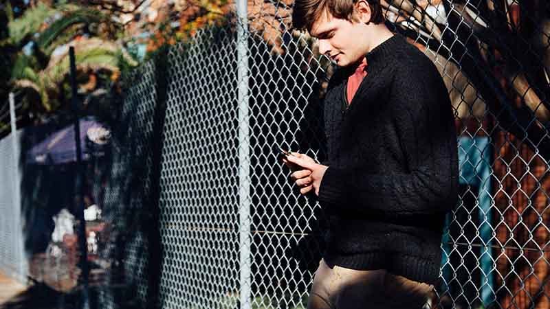 Young male leaning on fence using phone