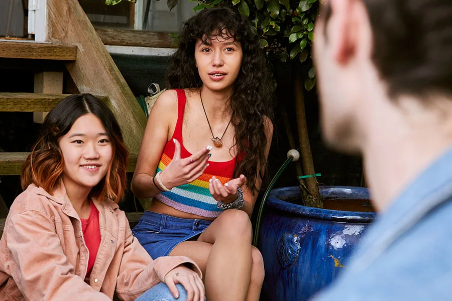 Two teenage girls sitting on steps facing a teenage boy. One of the girls is speaking passionately.