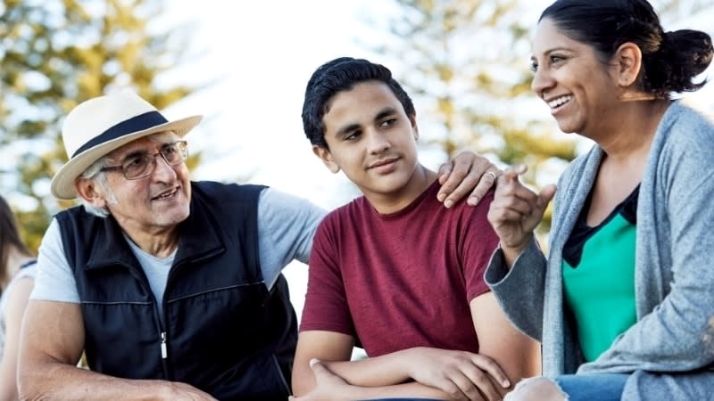 A young teenage boy smiles as he sits in the middle of his parents. His mother (right), has long dark hair in a ponytail and is wearing a green shirt and cardigan. His father (left), and his father, is wearing a dark vest and a white fedora hat, and has his arm around his son.