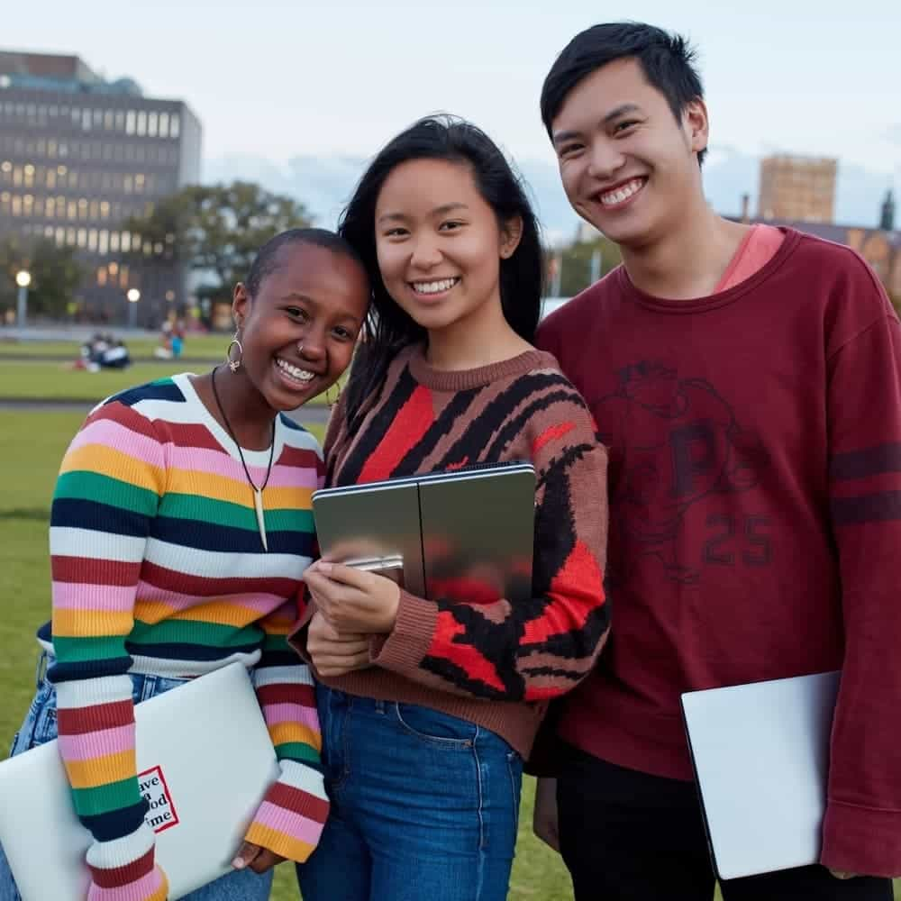 Image of three teenagers standing close together and smiling to the camera. The woman on the left has black skin and shaved hair. She is resting her head on the woman in the middle, who has tanned skin and long black hair. The man on the right has light skin and short black hair.