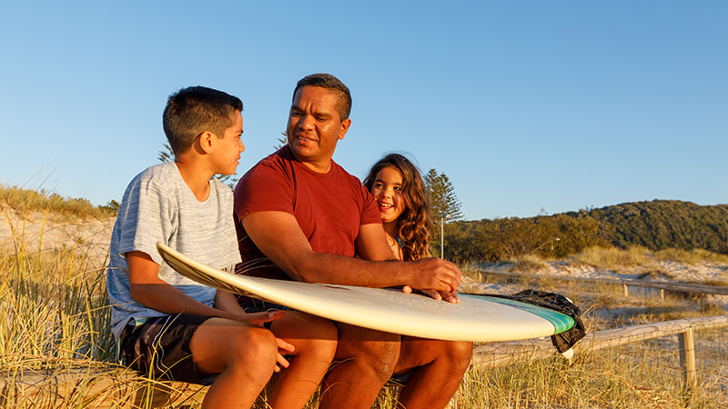 aboriginal dad and two teens talking holding a surfboard