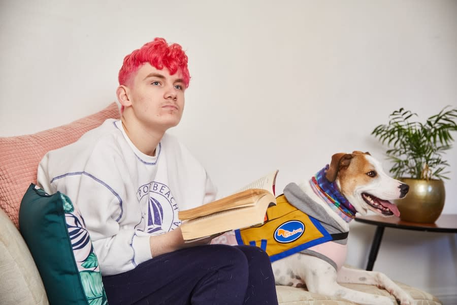 young person sitting on a couch next to their dog reading a book staring into the distance