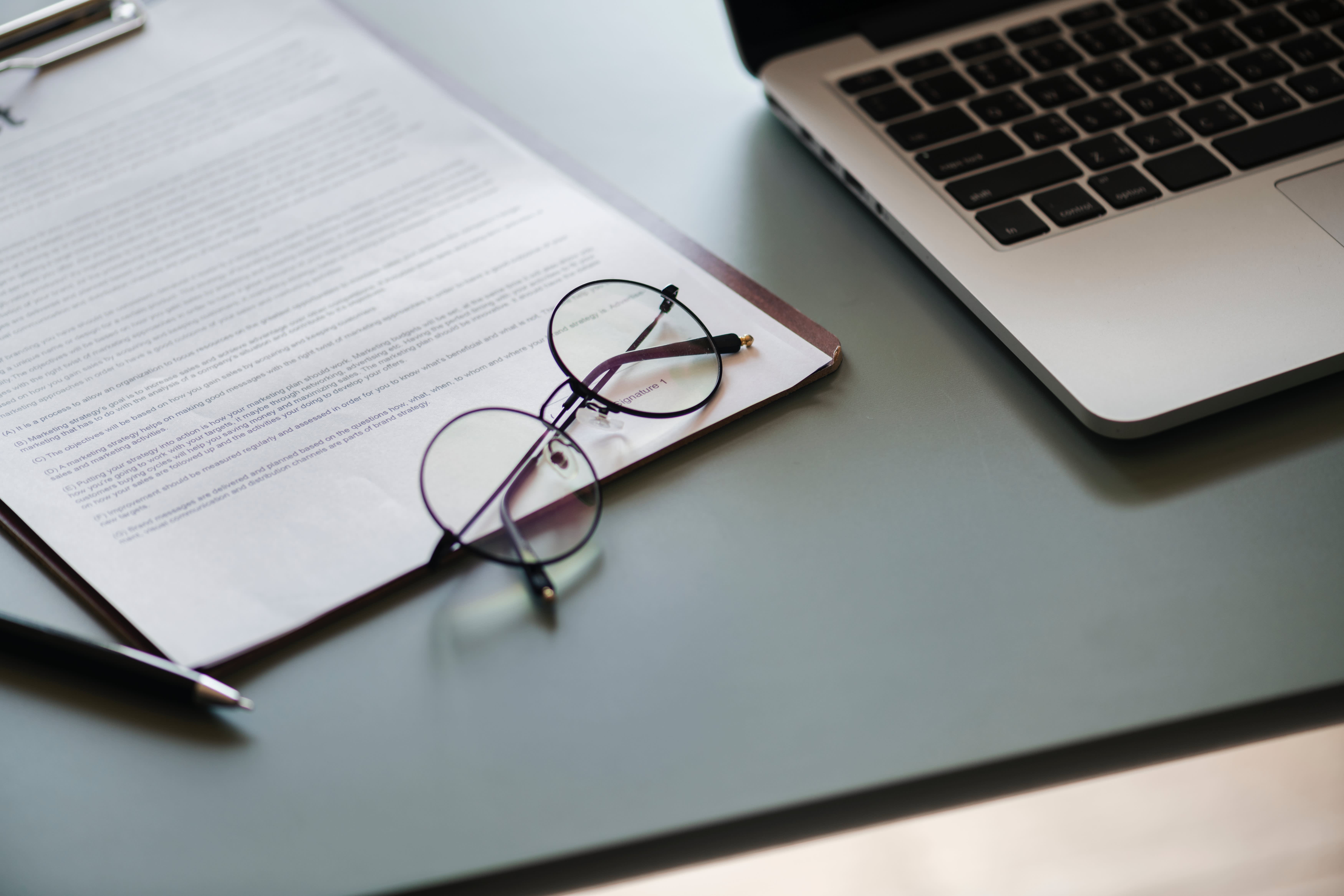 Paperwork with glasses and pen next to computer on desk