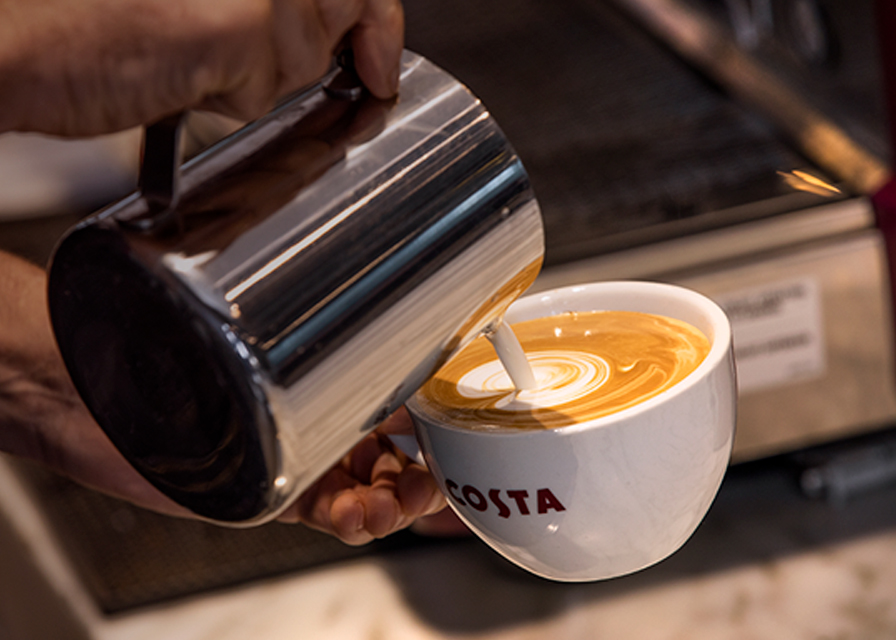 Picture of a Costa coffee Barista pouring steamed milk into a Costa Coffee Cup that has shots of espresso in it, to create a signature Flat White.