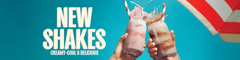 Two hands hold up strawberry and chocolate milkshakes in glass jars with striped straws, splashing at the top against a bright blue background. Large text on the left reads “New Shakes: Creamy-Cool & Delicious.”