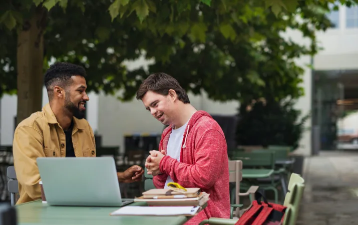 A male social worker of colour chatting to a man with Down syndrome outdoors over a laptop