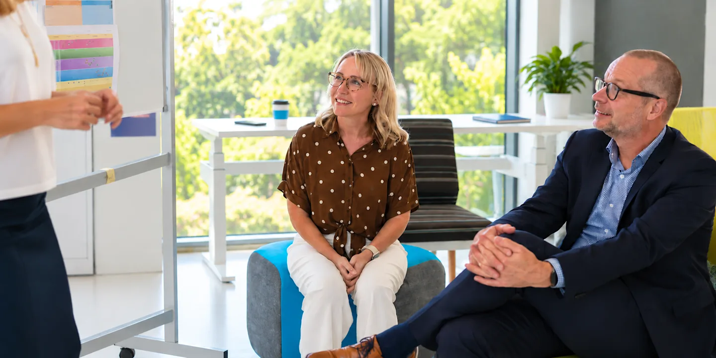 University professionals listen to a presenter during a meeting in a bright office with a leafy outlook.
