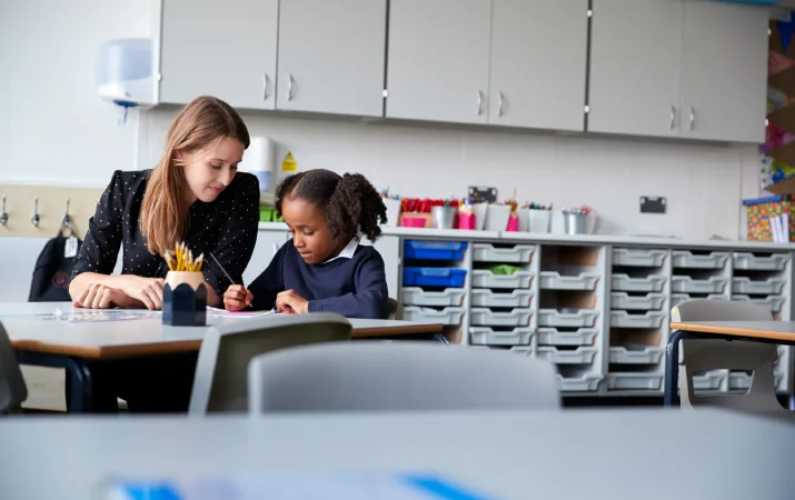 A young female primary school teacher working one-on-one with a pupil in the classroom