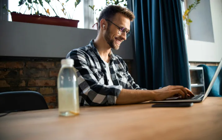 A law student sits at a table smiling while studying with classmates via his laptop at home