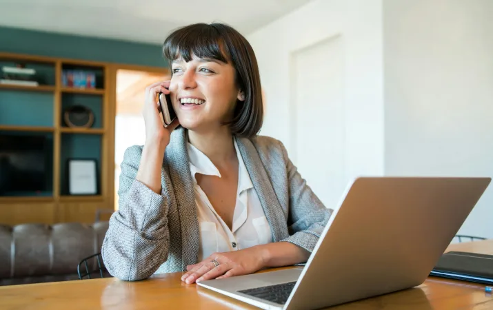 A woman using a laptop while speaking on the phone at home.