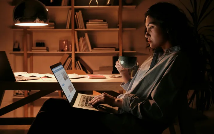 Woman browsing a website on her laptop while enjoying a cup of tea in a warmly lit home study.