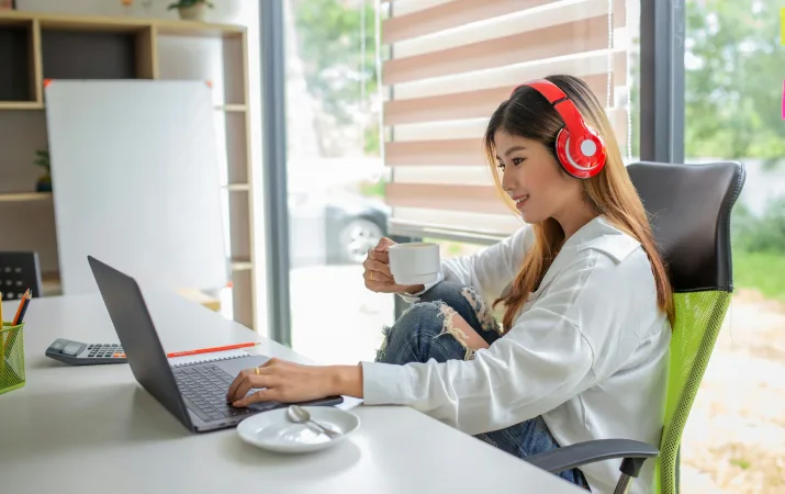 Person wearing red headphones uses a laptop at a desk beside a window.