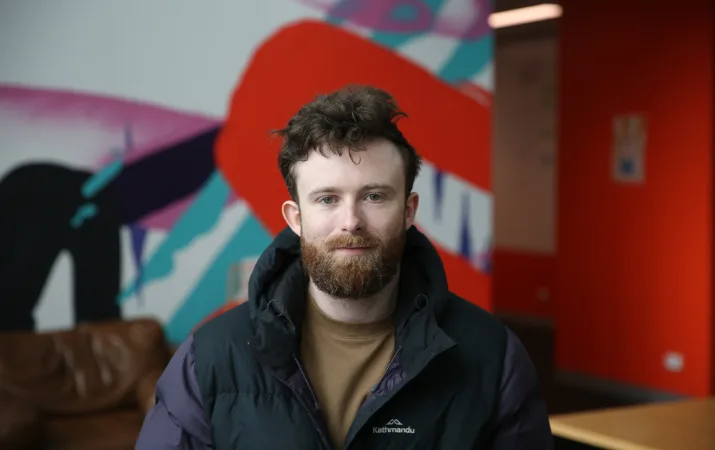 Young man with beard who looks calm and confident as he stands in front of office mural