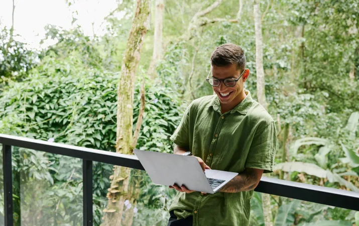 A young man standing on a balcony overlooking a beautiful regional forest grinning down at his laptop