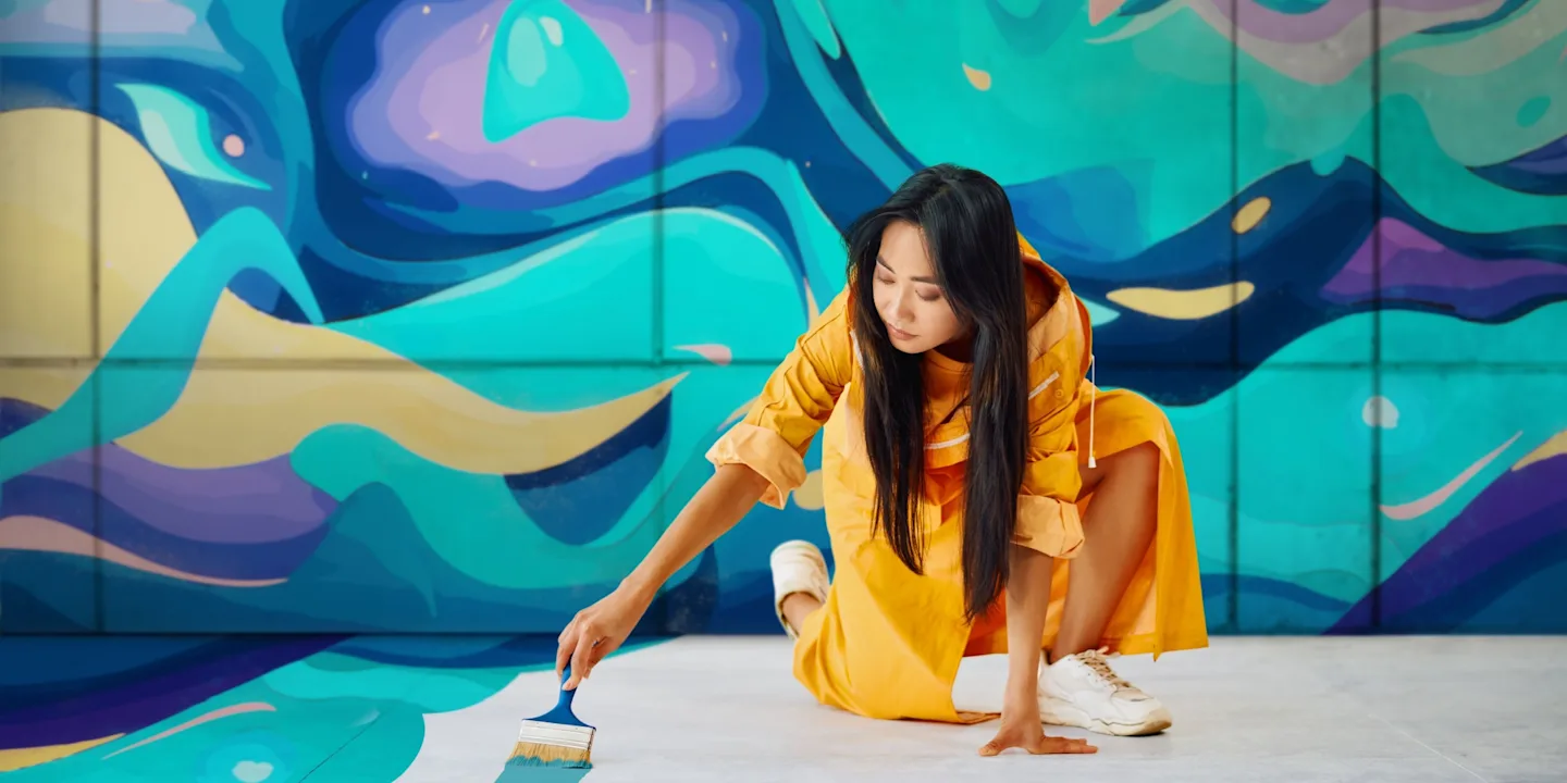 An artist painting a colourful abstract mural on a wall and floor with a paintbrush.