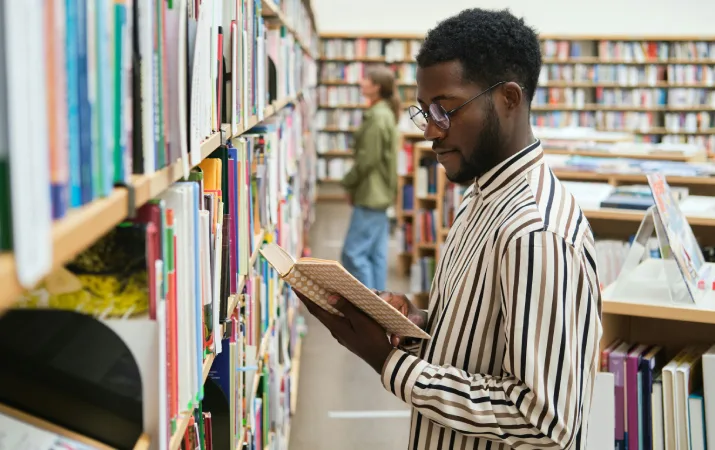 University student wearing glasses and a striped shirt reading a book beside bookshelves in a library.