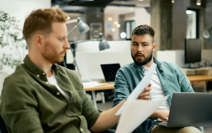 Two colleagues review printed reports together at a desk in an office.