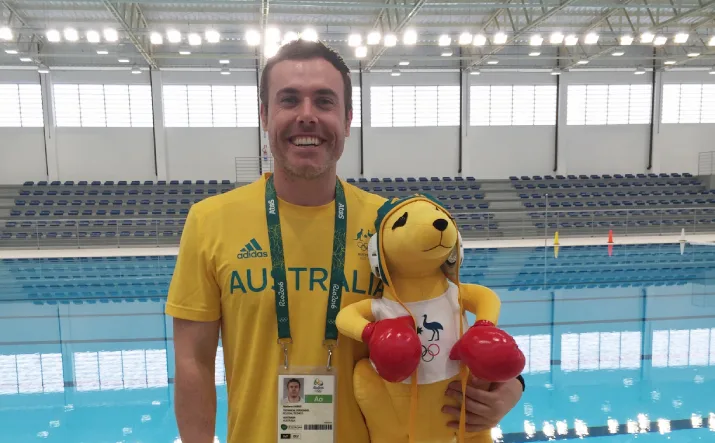 Mat Harris, Performance Science Assistant Manager at the VIS, standing in front of a swimming pool at Rio Olympics, holding a plush boxing kangaroo toy mascot.