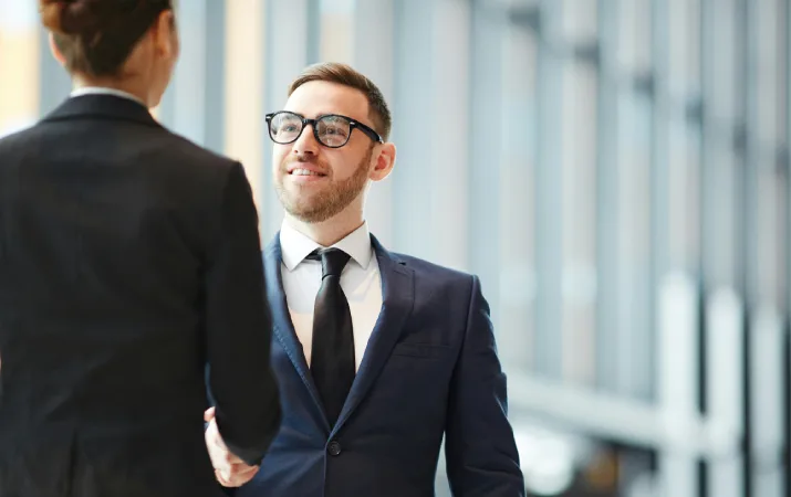 Smiling man in a suit and glasses shaking hands with another professional in a modern office.