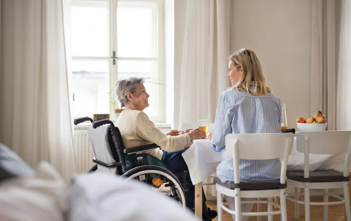 Elderly woman in wheelchair smiling and eating lunch with a younger woman