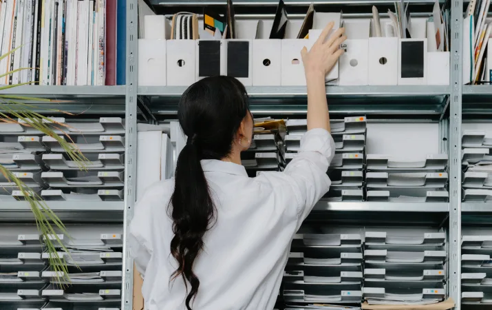 A young female paralegal facing a large shelf of case files, reaching for a binder