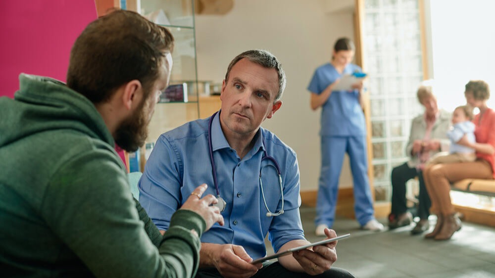 A bearded man in a green hooded sweatshirt is seen speaking with a doctor. The doctor's expression is strained. Behind them, a family speak with another medical professional.