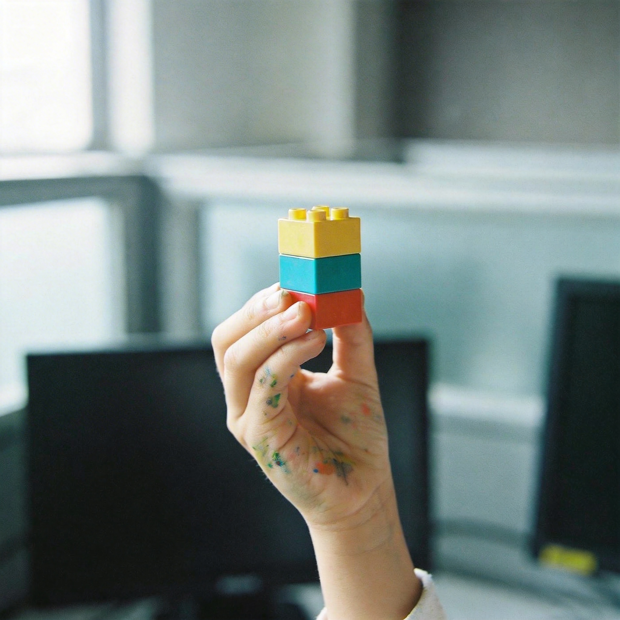 Child's hand with building block against blurred office background, representing lost creativity.