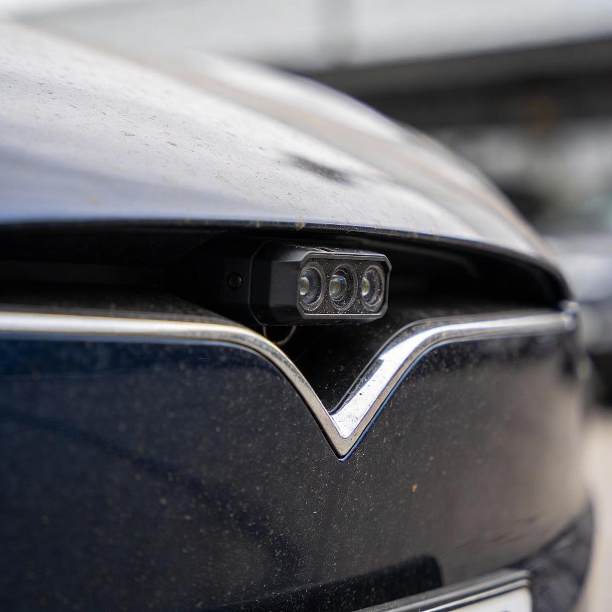 Close-up of a Tesla's front grille and sensor array, showing subtle dust, against a blurred urban background.