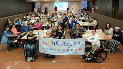A group of wheelchair users seem to be in a hall, looking at the camera while 2 of them holding a sign with the word WheeLog on it