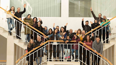 A group photo of males and females standing at the stairs smiling and waving while looking at the camera
