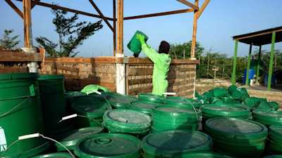 A man wearing light green uniform surrounded by dark green-colored barrel emptying dirt from a barrel
