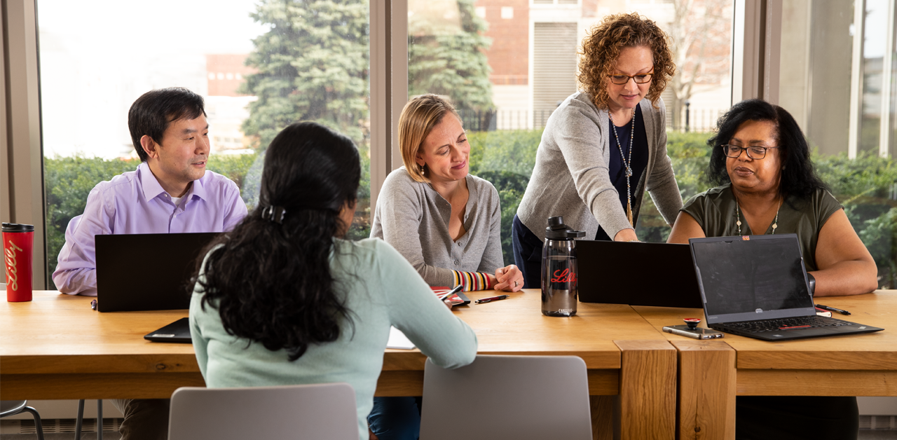 A group of five people working at a table in front of a large window. One women, who is wearing a grey cardigan, is leaning over the shoulder of another and pointing at somenthing on the laptop screen in front of her.