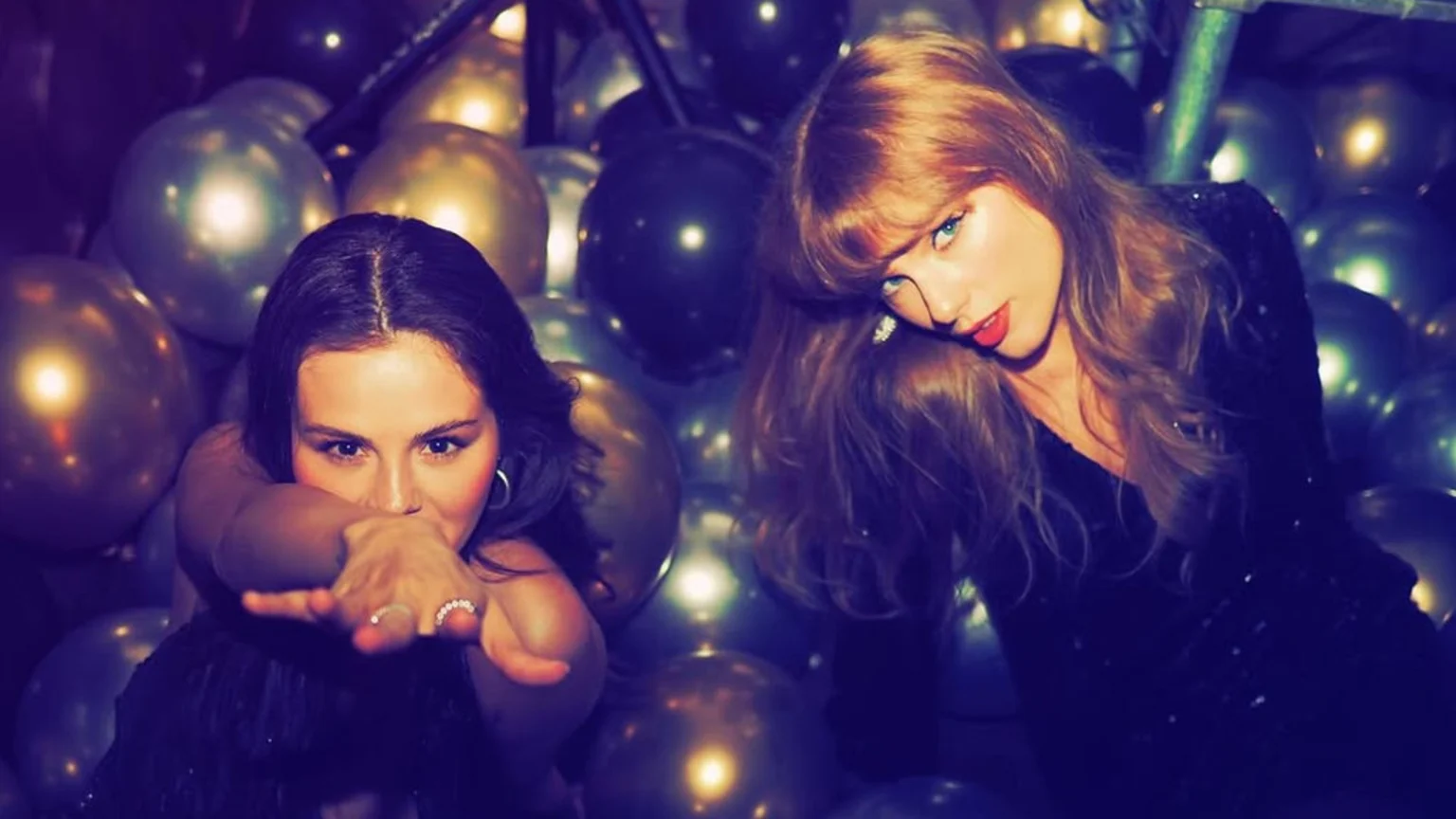 Selena Gomez and Taylor Swift, both in dark outfits, pose for the camera surrounded by shiny gold, silver, and dark balloons; Gomez's arms are crossed, and Swift leans forward with red lipstick.