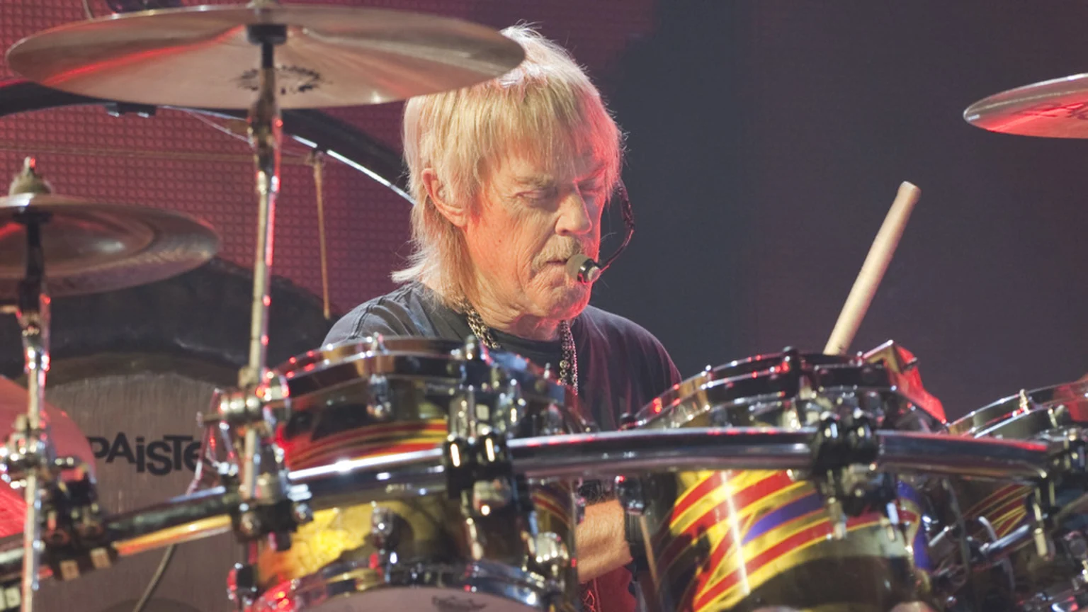 A male drummer with blonde hair and a mustache, wearing a headset microphone, plays a drum kit with colorful striped designs.