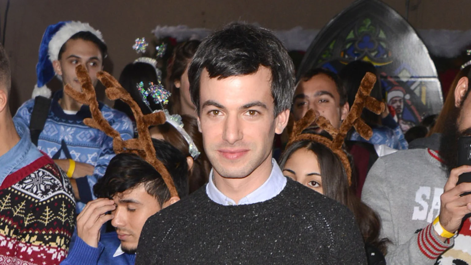 Nathan Fielder, a man with short dark hair, smiles at the camera while standing in a crowd of people, some wearing festive holiday headwear like reindeer antlers and a blue Santa hat.