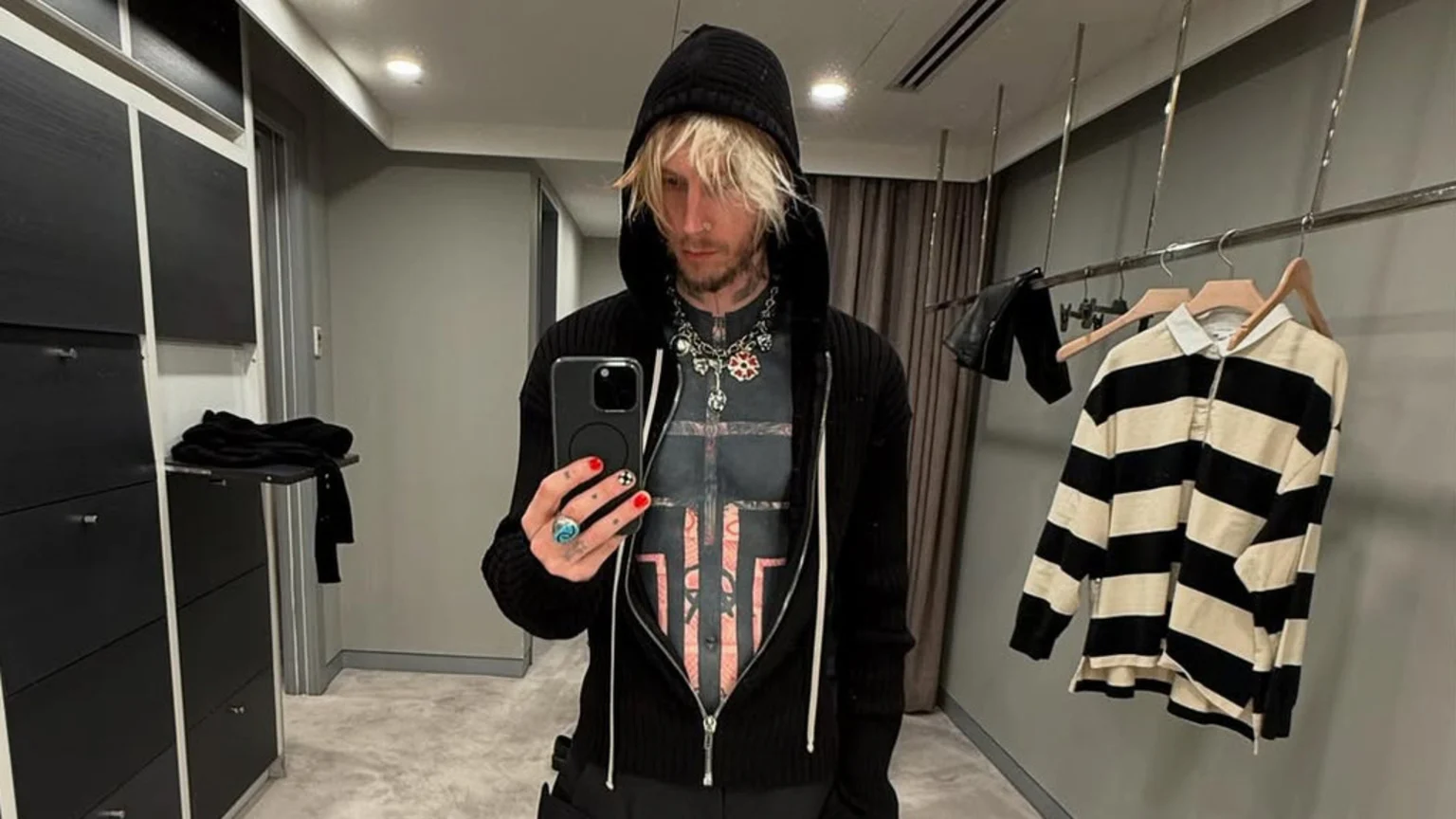 Machine Gun Kelly takes a mirror selfie in a black hoodie and graphic tee, showing his blonde hair, necklaces, painted nails, and a striped shirt on a rack behind him.