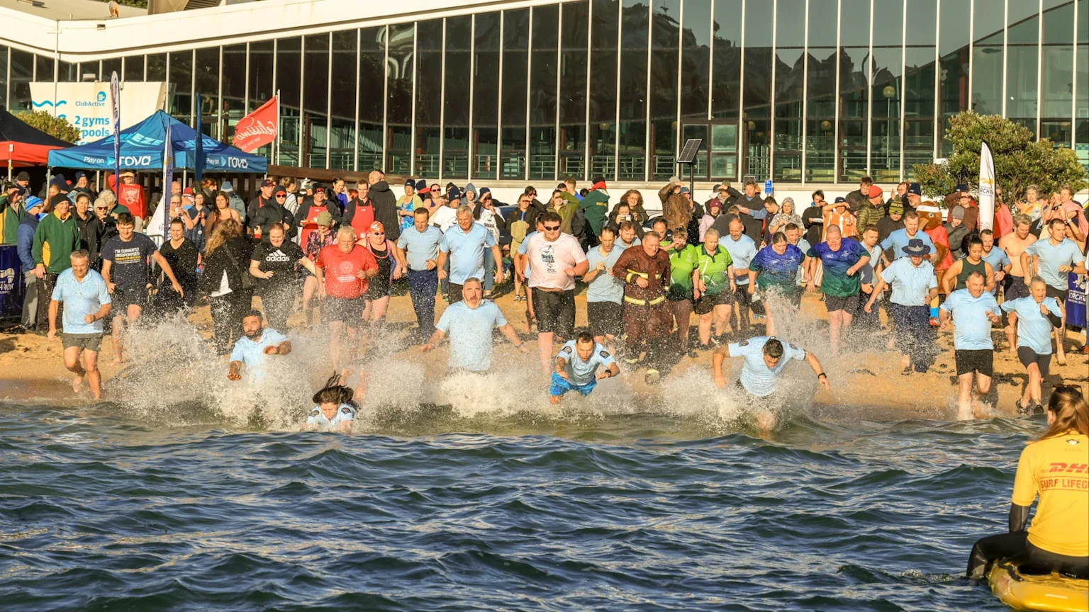 A large group of people enthusiastically run and splash into the water from a sandy beach, with a modern building and spectators visible in the background.