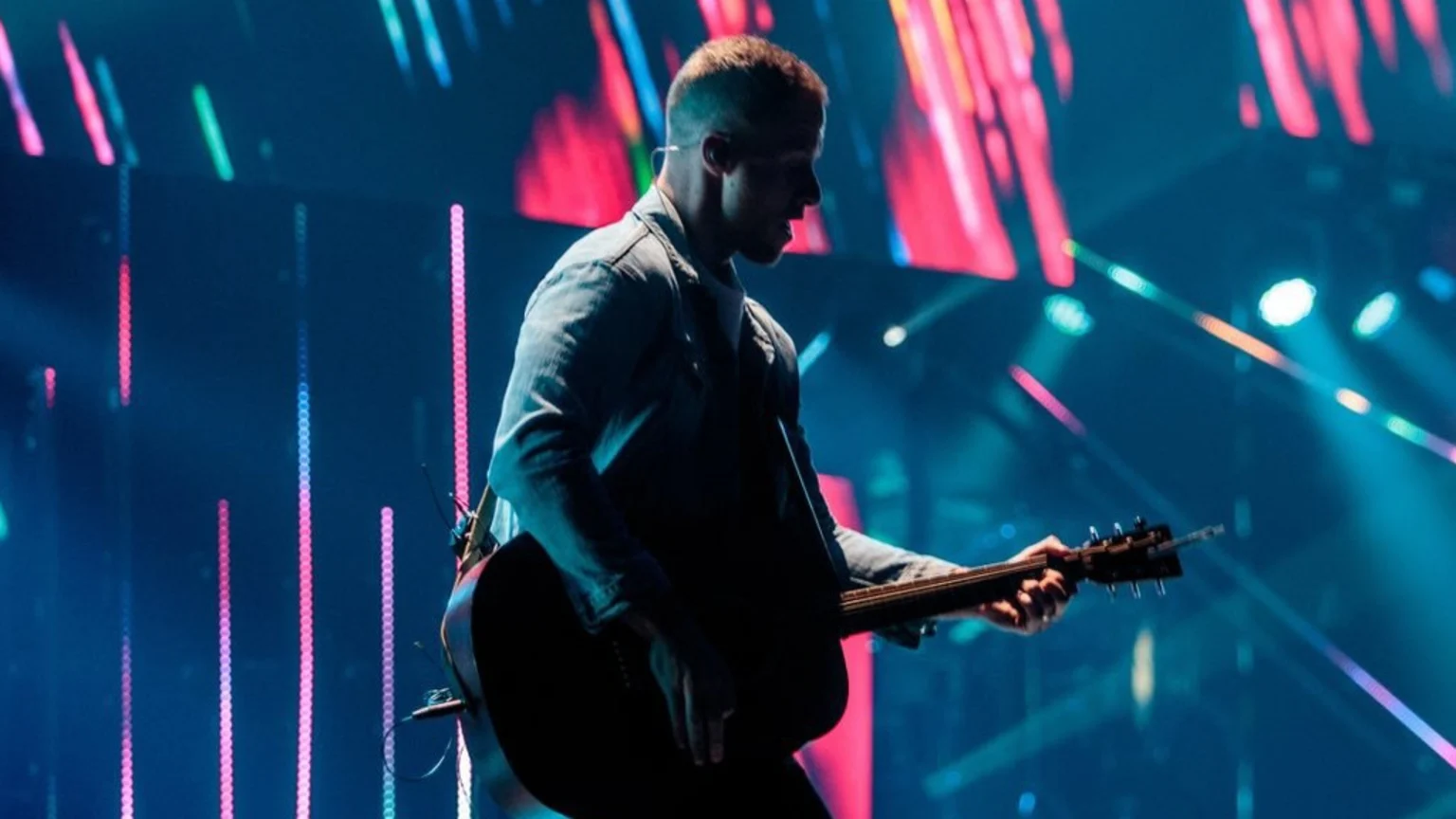 A man in profile, wearing a jacket, plays an acoustic guitar on a stage, silhouetted against vibrant blue, pink, and red stage lights.
