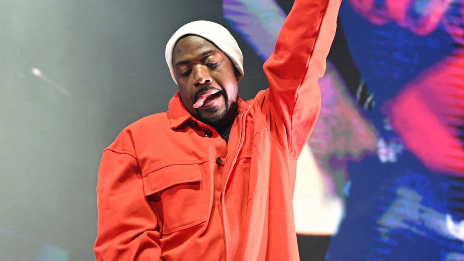 A man with a goatee, wearing a white beanie and orange shirt, sticks his tongue out and raises his left arm, against a blurry background of gray and colorful lights.