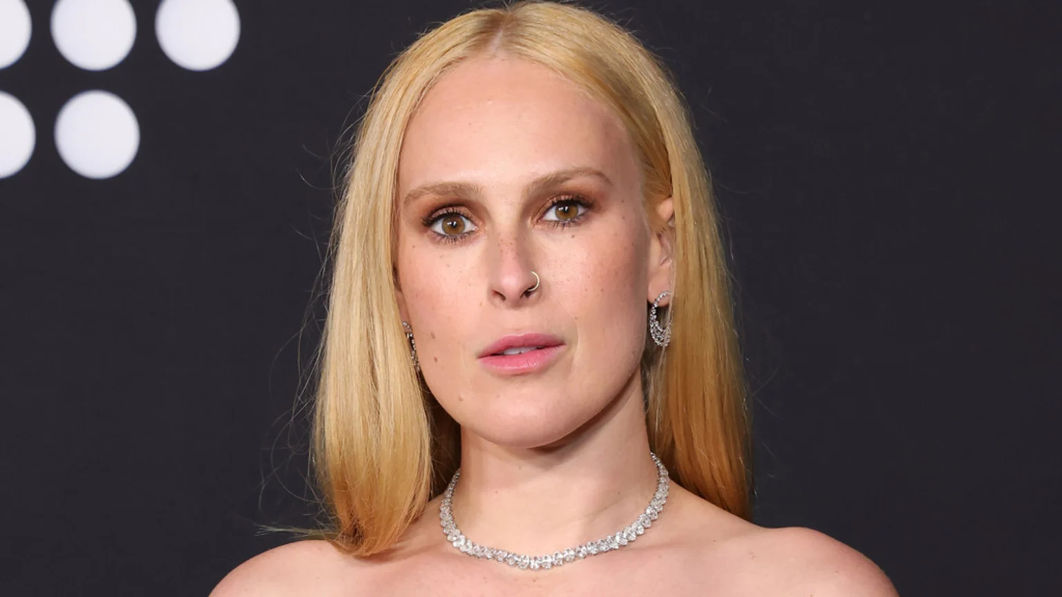 A blonde woman, Rumer Willis, with a nose ring and freckles, looks directly at the camera, wearing a diamond necklace and earrings against a dark background with faint white circles.