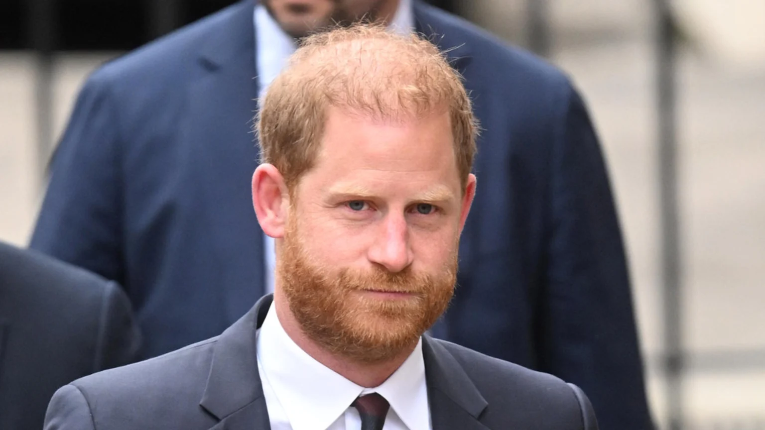 Prince Harry, a man with ginger hair and beard, wearing a dark suit and white shirt, looks directly at the camera with a neutral expression, with blurred figures in the background.