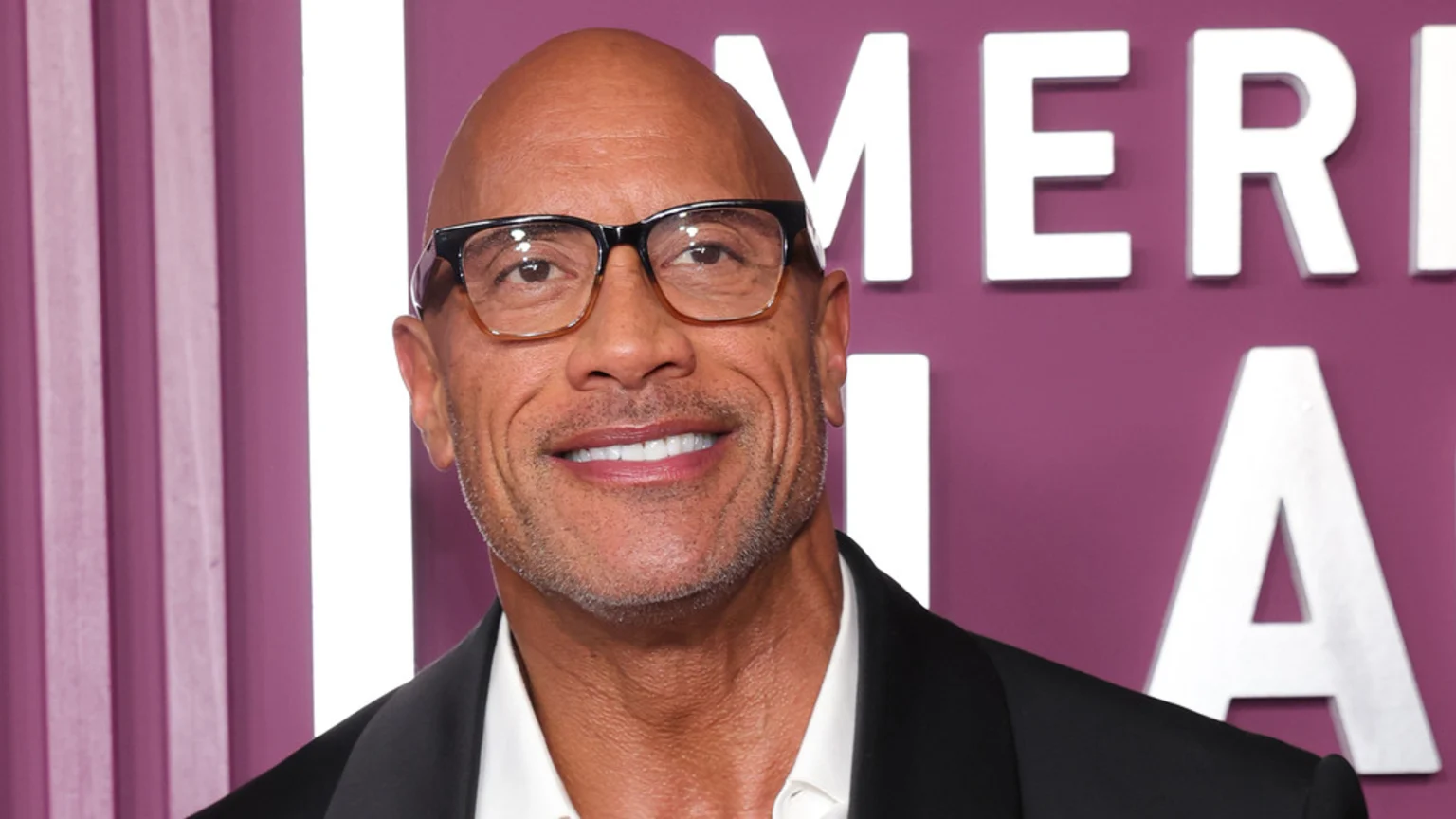 Close-up of Dwayne Johnson smiling and wearing glasses, a dark suit jacket, and a white shirt, with blurry white letters on a purple wall in the background.