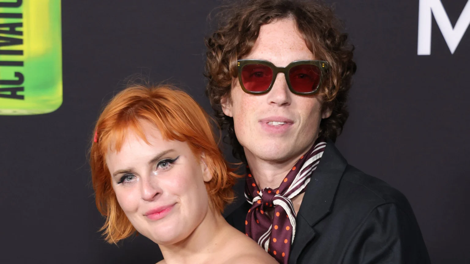 A woman with reddish-orange hair and winged eyeliner smiles beside a man with curly hair, red-tinted sunglasses, and a striped, polka-dotted scarf.