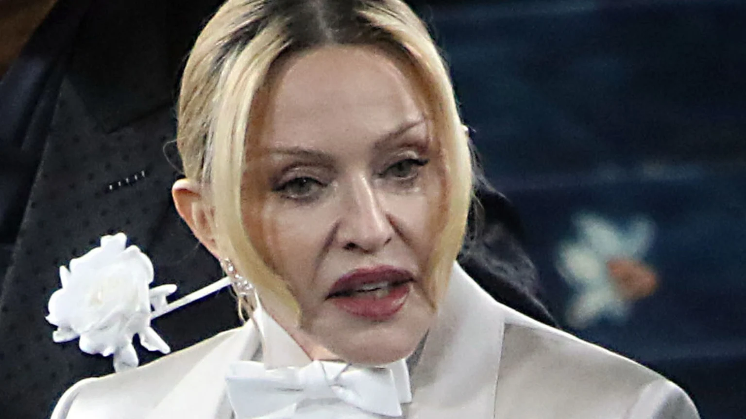 A close-up shot shows a woman with blonde hair and light eyes, wearing a light-colored suit, a white bow tie, and dark red lipstick. A white rose is pinned to a dark, subtly patterned lapel next to her.