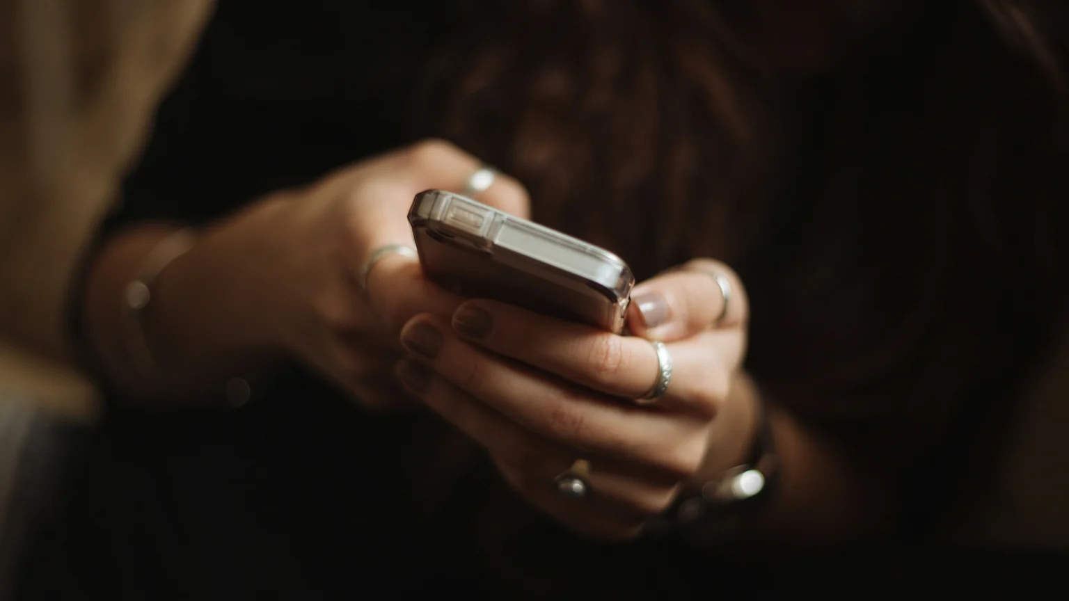 A close-up shows a person's hands holding a smartphone with a clear case, with rings on their fingers and painted nails, against a dark, blurred background.