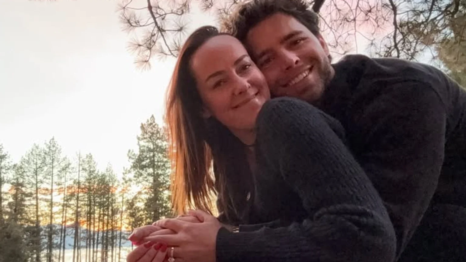 A smiling couple embraces outdoors during a golden sunset. The woman, wearing a dark long-sleeved sweater with a visible silver engagement ring, stands in front; the man stands behind her in a matching sweater. A forest of tall pine trees and distant snow-capped mountains form the scenic backdrop beneath a vibrant golden-orange sky.
