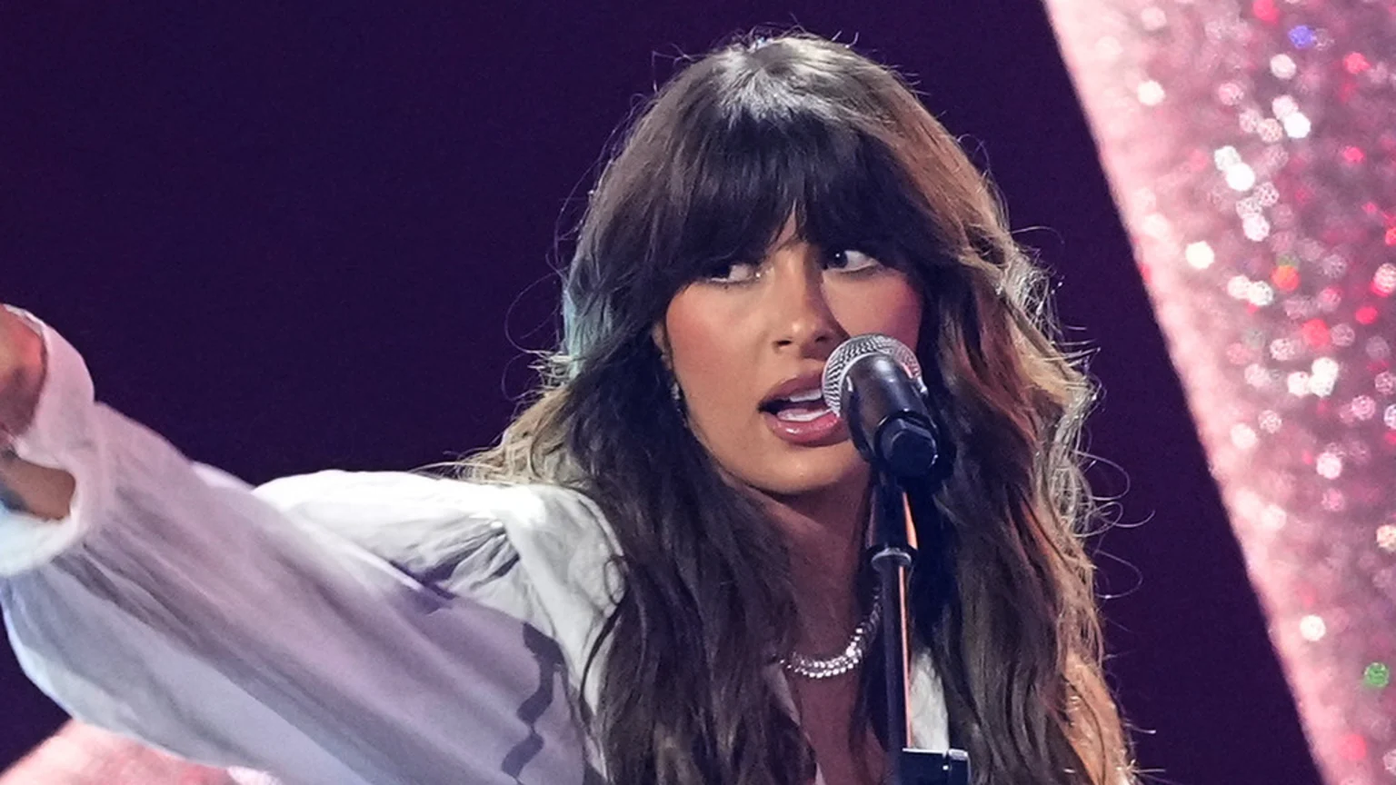 Ella Langley, a woman with long dark hair and bangs, sings into a microphone with her mouth open and left arm raised, wearing a light blouse and necklace, against a sparkling pink and dark background.