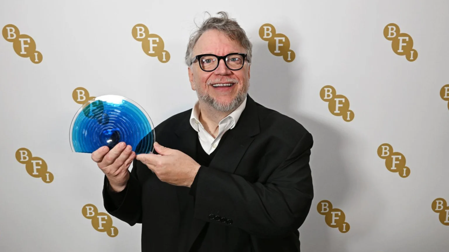 Guillermo del Toro, smiling in glasses and a suit, holds a blue, circular award while gesturing towards it, against a white BFI logo backdrop.